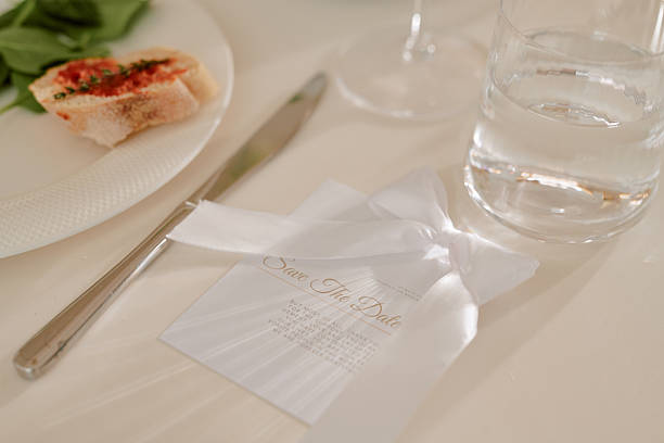 Closeup of wedding invitation card with ribbon placed on table near glass of water, knife, and plate with bread and greens, capturing details of wedding celebration setup