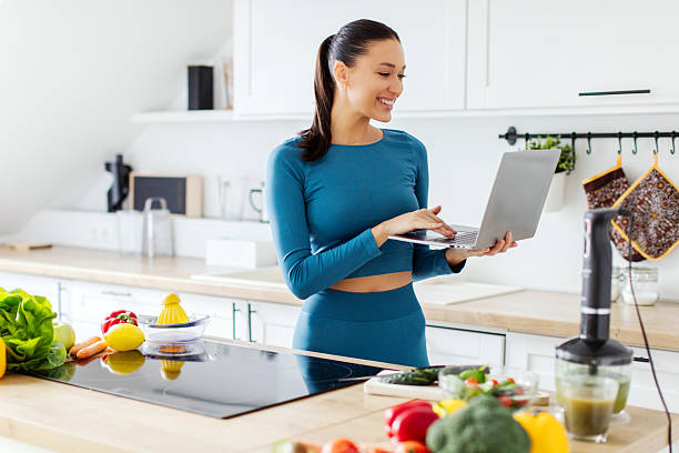 Happy fit lady in sportswear using laptop browsing nutritious recipes for slimming, planning menu online standing in kitchen. Weight loss concept