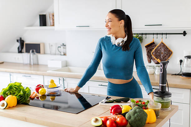 istock Young sporty lady preparing fresh vegetable salad in light modern kitchen interior, looking aside at free copy space and smiling 2219515955