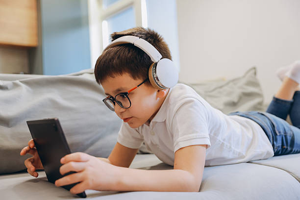 istock Young boy with headphones and eyeglasses using tablet while lying on sofa at home 2219519044