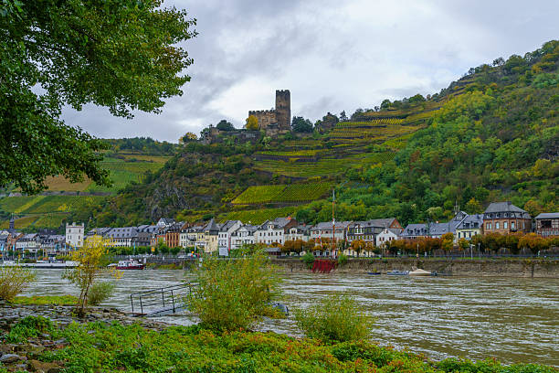 The black, red and yellow German flag flies off the stern of a passanger ship while passing Kaub, Germany and Gutenfels Castle in the Upper Middle Rhine Valley in Rhineland-Palatinate during a sightseeing pleasure cruise on the Rhine River. In the background, lush, wooded rolling hills on the north side of the river can be seen descending down to the medieval riverfront town and wtaer's edge.