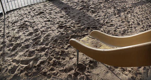 A bright yellow plastic playground slide sits in a sandy playground area with textured footprints and patterns scattered across the sand surface. The slide's curved form creates shadows in the natural daylight illuminating the play area.\nPhoto is 1.25x anamorphic