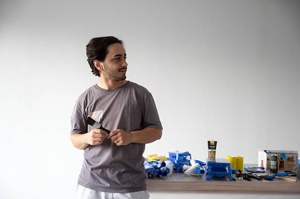 Young adult painter holding paintbrush and looking away in empty room with painting equipment