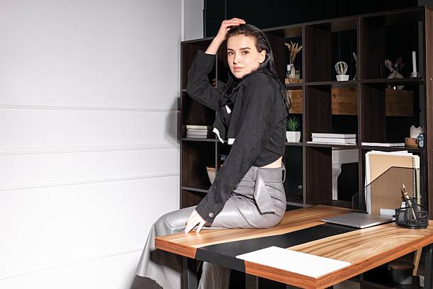 Elegantly dressed individual sits on a wooden desk in a sleek office space, exuding confidence and style while engaging with the camera.