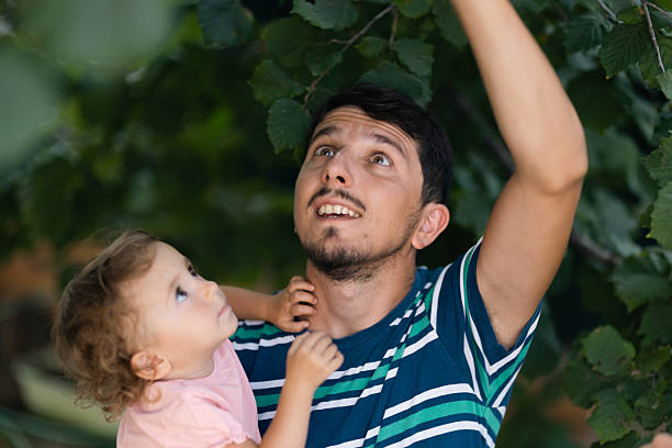 Father holding his child as they observe and interact with fresh tree leaves in an outdoor setting. The family shares a bonding moment surrounded by nature's beauty and bright green foliage.