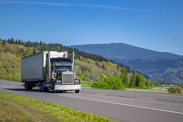 istock Black classic big rig semi truck tractor transporting cargo in refrigerator semi trailer running on the flat highway road with green hills on the side 2219567449