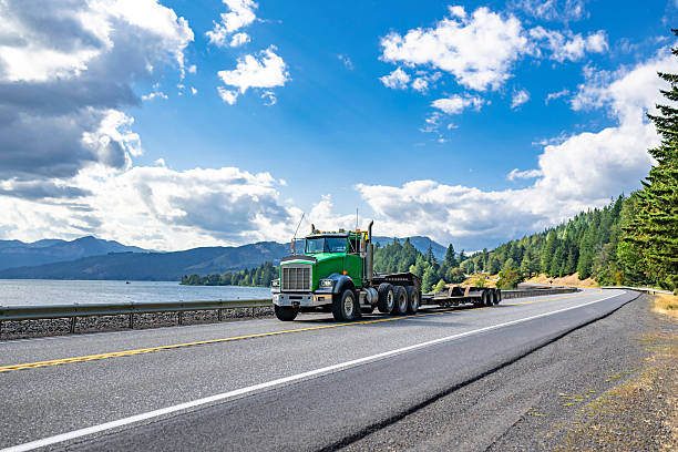 istock Green powerful big rig semi truck tractor transporting empty heavy duty step deck semi trailer running on the highway road along the Columbia river in Columbia Gorge area 2219567931