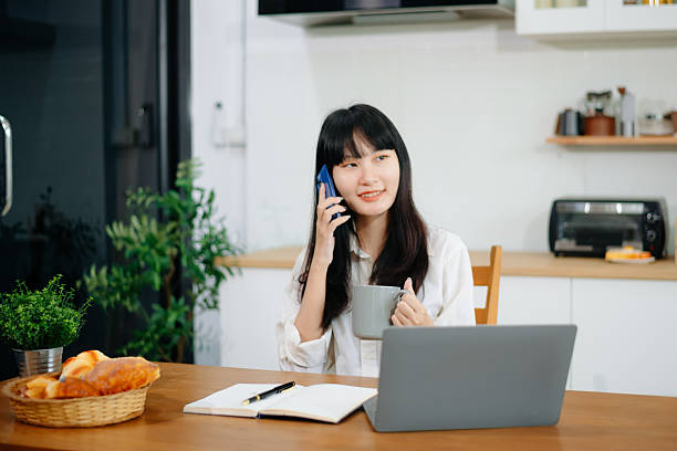 istock Asian woman working from home while talking on phone in kitchen. Perfect for remote work 2219573597