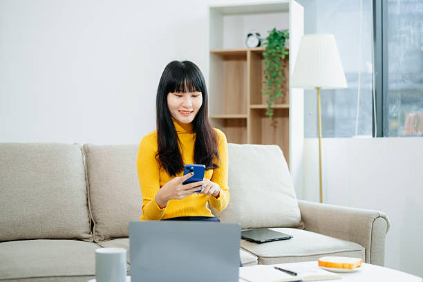 istock Asian woman sitting on sofa using tablet and drinking coffee while working from home. Great for remote work, digital lifestyle, relaxation 2219573607
