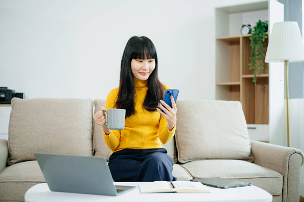 istock Asian woman sitting on sofa using tablet and drinking coffee while working from home. Great for remote work, digital lifestyle, relaxation 2219573610