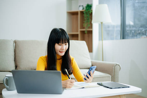 sian woman writing notes while using phone and working remotely with laptop. Ideal for themes of freelance, productivity, multitasking