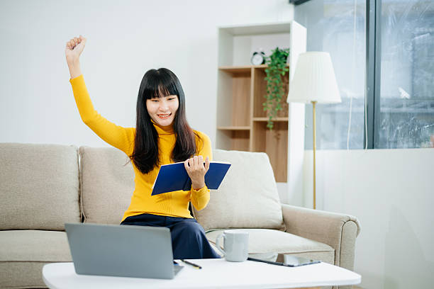 istock sian woman writing notes while using phone and working remotely with laptop. Ideal for themes of freelance, productivity, multitasking 2219573613