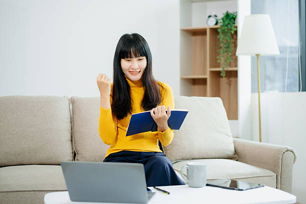 sian woman writing notes while using phone and working remotely with laptop. Ideal for themes of freelance, productivity, multitasking