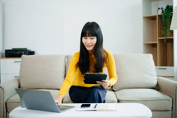 istock Asian woman sitting on sofa using tablet and drinking coffee while working from home. Great for remote work, digital lifestyle, relaxation 2219573617