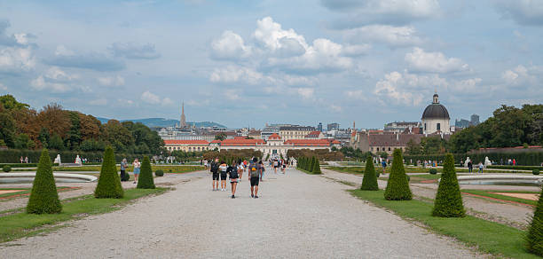 Vienna, Austria - August 31, 2022: A wide stone walkway flanked by sculpted shrubs and flowerbeds leads through the historic gardens of Belvedere Palace. Tourists stroll along the path, taking in the classic landscaping.