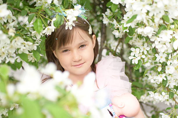 Portrait of a beautiful little girl in the bushes of a blooming apple tree. A doll in his hands.