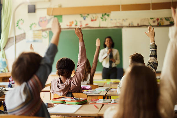 istock Back view of students raising their hands to answer the teacher's question on a class. 2219587556