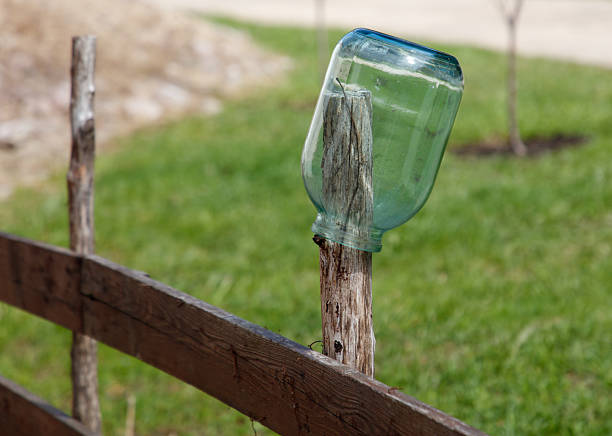 A green jar is sitting on a wooden post. The jar is upside down and he is broken. The scene is set in a grassy area with a fence in the background