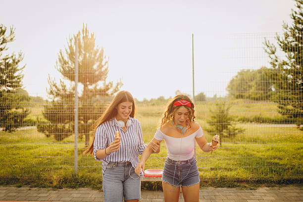 Two young cheerful female friends enjoying ice-cream while roller skating on outdoor sports court during the sunny day