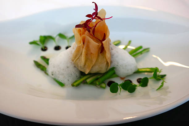 This exquisitely plated dish features a delicate, golden dumpling, perfectly enclosed and artfully presented as a culinary gift. It rests gracefully atop a bed of vibrant green asparagus spears, each cooked to tender-crisp perfection. A ethereal foam adds a cloud-like texture, while dark, glistening droplets of sauce and fresh microgreens provide elegant accents, promising a sophisticated and utterly delicious experience.