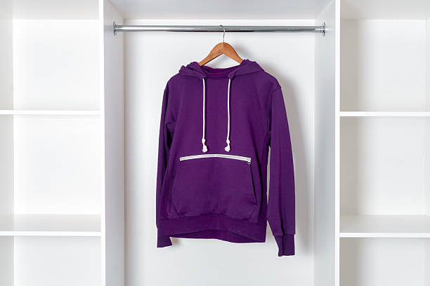 Purple hoodie with white drawstrings and zipper pocket hanging on a wooden hanger inside a modern white wardrobe. stock photo