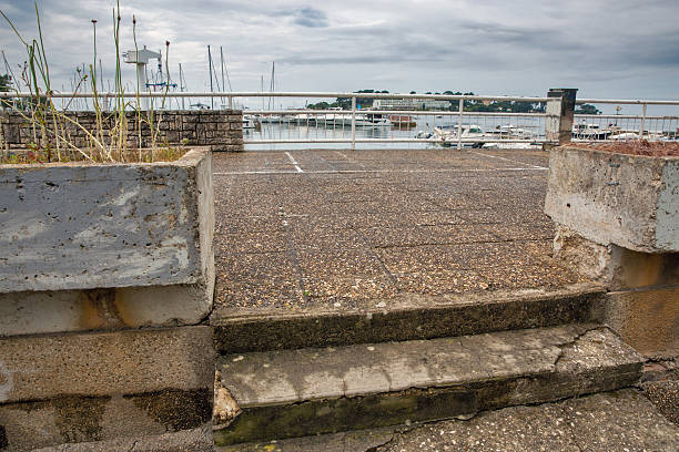 Weathered concrete stairway leads to a marina promenade with boats and yachts in the background under a cloudy sky. A scene of coastal urban decay and charm in Porec, Istria, Croatia.