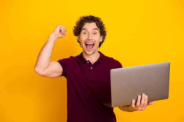 istock Excited man holding laptop on vibrant yellow background with cheerful expression celebrating success and achievement 2219607713