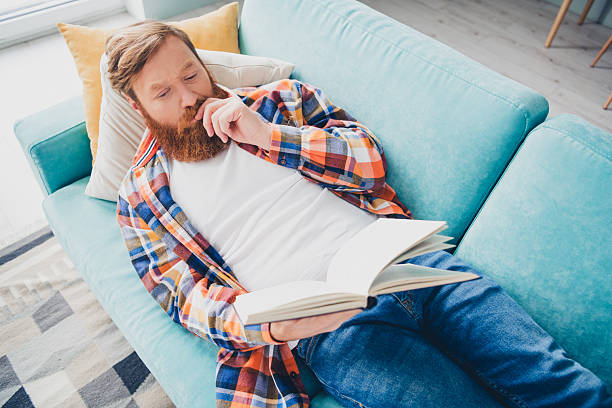 istock Relaxed young man with red beard and checkered shirt reading a book on comfortable couch in interior living room 2219607976