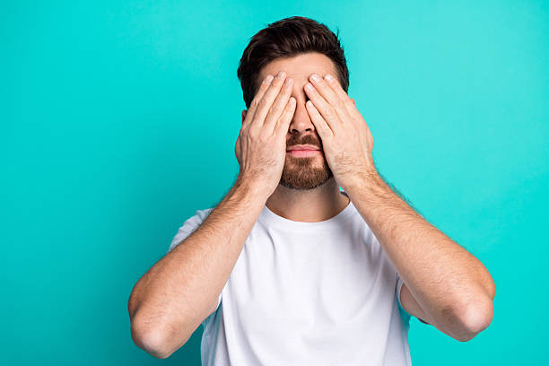 istock Young man covering his eyes with hands on a teal background, expressing playfulness and curiosity in a casual style 2219608577