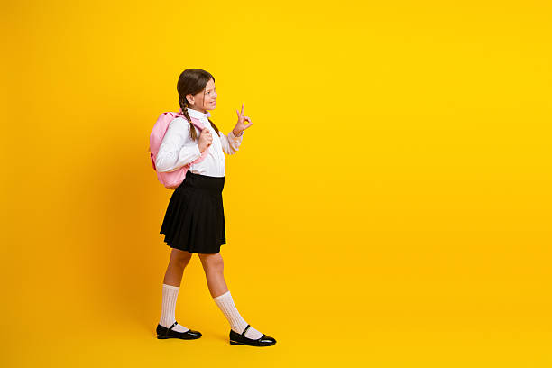 istock Confident young schoolgirl in uniform with a pink backpack posing on a yellow background, enjoying back-to-school season 2219612603