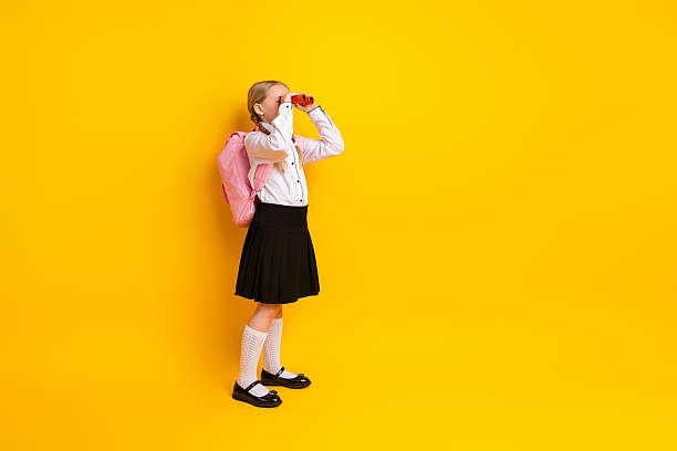 istock Smiling schoolgirl with pink backpack looking through binoculars against yellow background showcasing education and exploration 2219615528