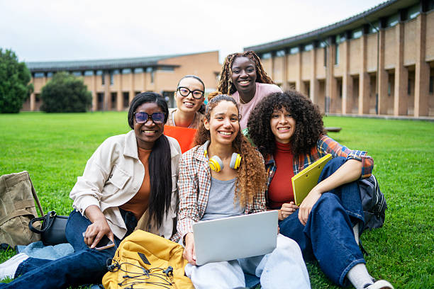 Diverse female students collaborating, studying together on green campus lawn, sharing cheerful moment