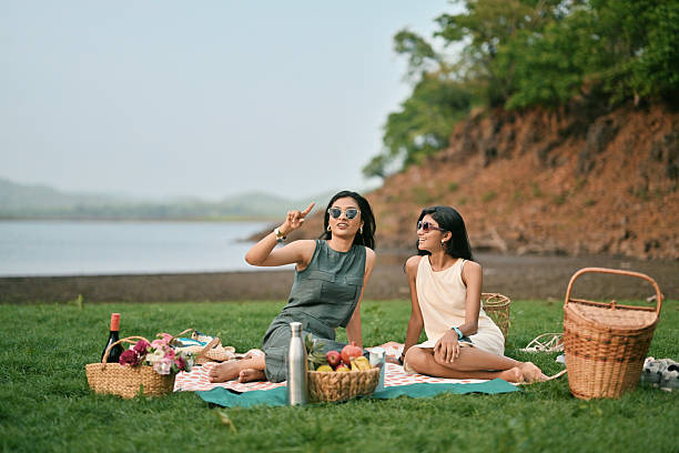 Two girls sit on a picnic mat near a lake, surrounded by baskets of fruit and flowers. One of them flashes a peace sign while the other smiles, capturing a lighthearted moment in a serene natural setting.