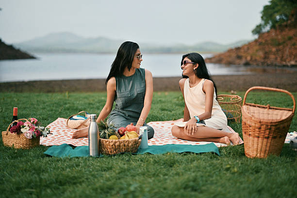 Two girls sit on a picnic mat near a lake, surrounded by baskets of fruit and flowers. One of them flashes a peace sign while the other smiles, capturing a lighthearted moment in a serene natural setting.