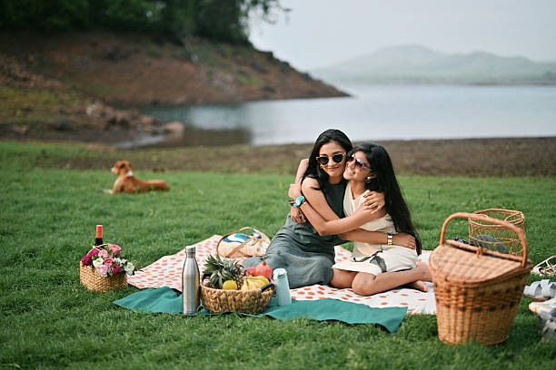 Two sisters sitting on a blanket at a lakeside picnic, hugging and smiling at the camera. Picnic baskets, food, and drinks are arranged around them on the grass. A dog is lying in the background near the lake.