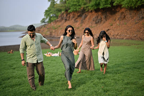 A family walks barefoot on grass, holding hands and enjoying the outdoors. The lake and hills form a peaceful backdrop with picnic items behind them.