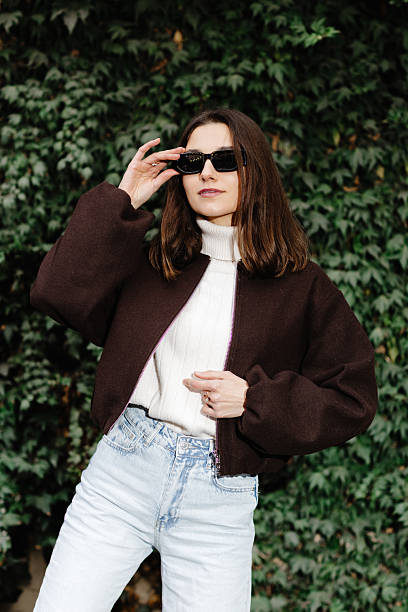 istock Stylish woman adjusting sunglasses while posing near ivy wall 2219624000