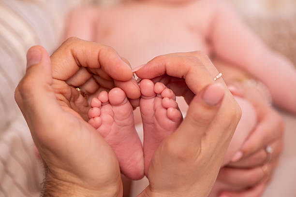 A heartwarming close-up image featuring the tiny feet of a newborn baby cradled and gently framed by the hands of two adults, likely parents. The fingers create a heart-like shape around the baby's soles, symbolizing love, care, and family unity. The soft lighting and shallow depth of field emphasize tenderness and emotion.