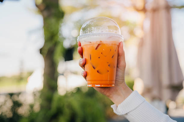 istock Thai Iced Tea. Refreshing Orange Iced Beverage Served in Clear Plastic Cup with Lid Held by Hand Against Bright Sunny Background 2219631644