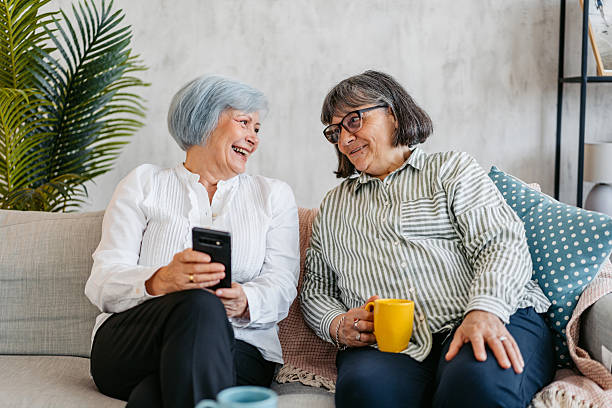 istock Two Senior Female Friends Drinking Coffee While Using A Phone Together On The Sofa In The Living Room 2219634909