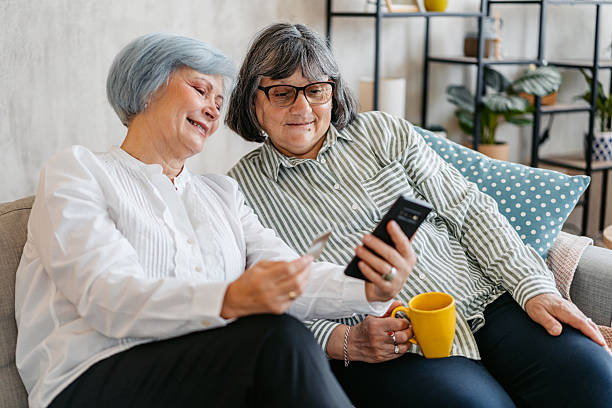 istock Two Senior Female Friends Drinking Coffee While Shopping Using A Phone Together On The Sofa In The Living Room 2219635161
