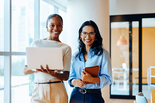 Two women smiling in an office holding laptop and notebook