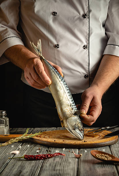 istock A skilled chef is holding a fresh mackerel above a wooden cutting board, surrounded by herbs and spices, showcasing culinary preparation in a cozy kitchen atmosphere 2219639923