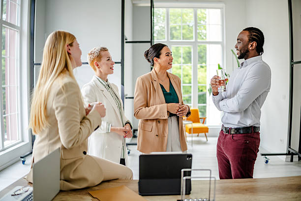 In a stylish office, a confident young black man engages with three female colleagues, sharing insights over drinks while fostering teamwork and collaboration.