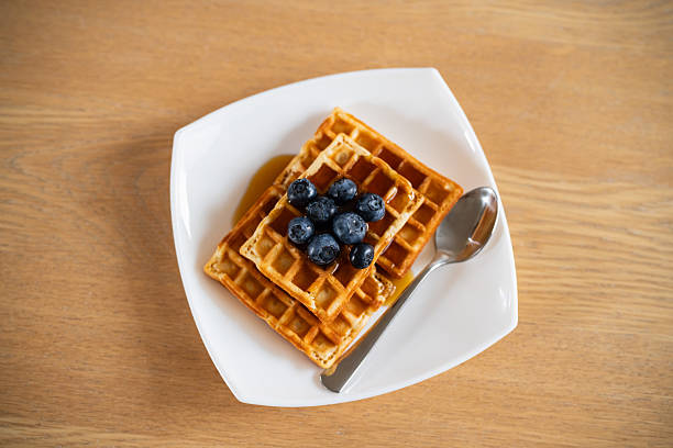 Delicious golden waffles served on a white plate, topped with fresh blueberries and maple syrup, alongside a silver spoon, creating a visually appealing and appetizing breakfast or brunch dish.