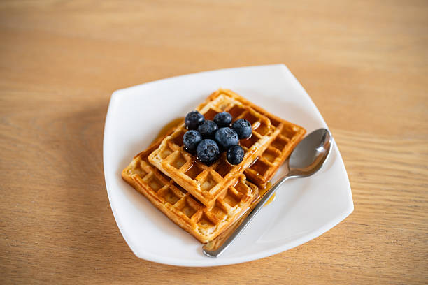 Freshly baked waffles topped with blueberries and syrup, served on a contemporary white plate with a spoon, creating a delightful breakfast setup perfect for food enthusiasts.