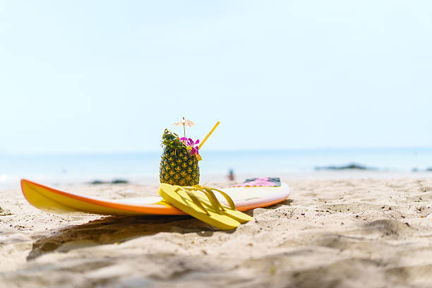 Beautiful decorated pineapple and flip-flops and surfboard left on the beach. Holidays, vacations, and summer vibes concept.