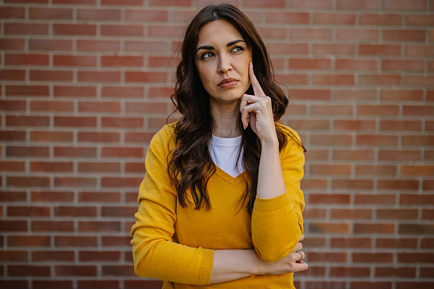 A thoughtful woman with her finger on her chin contemplates against a brick wall, depicting a moment of reflection, curiosity, and deep thought in an authentic manner.