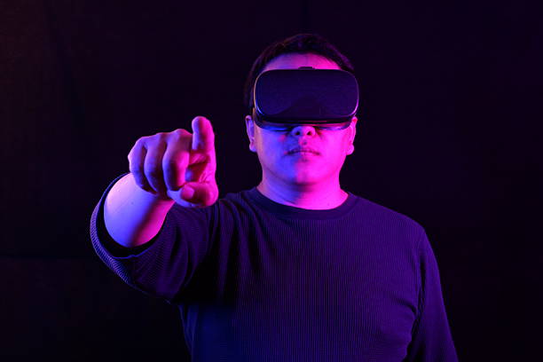 istock A person wearing a VR headset, pointing upwards with one finger as if interacting with a virtual screen, bathed in vibrant blue and pink neon light on a dark background. 2219660820