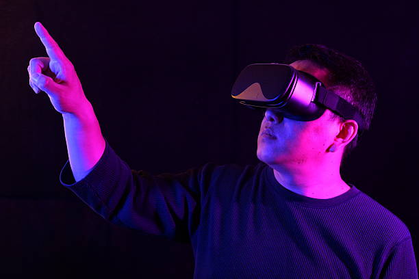 istock A person wearing a VR headset, pointing upwards with one finger as if interacting with a virtual screen, bathed in vibrant blue and pink neon light on a dark background. 2219660858